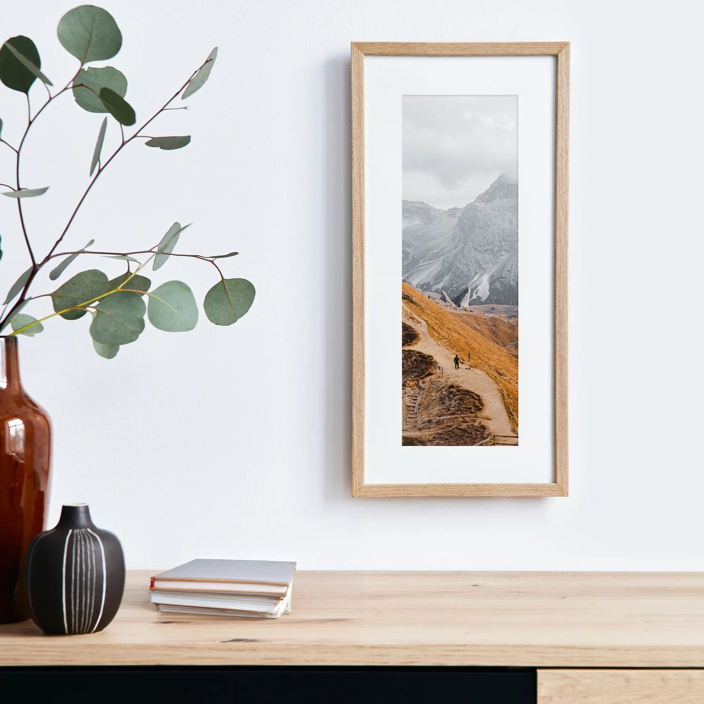 Vertical panoramic print of a winding mountain trail with a hiker and dramatic rocky peaks, displayed in a light wood frame above a wooden console table.
