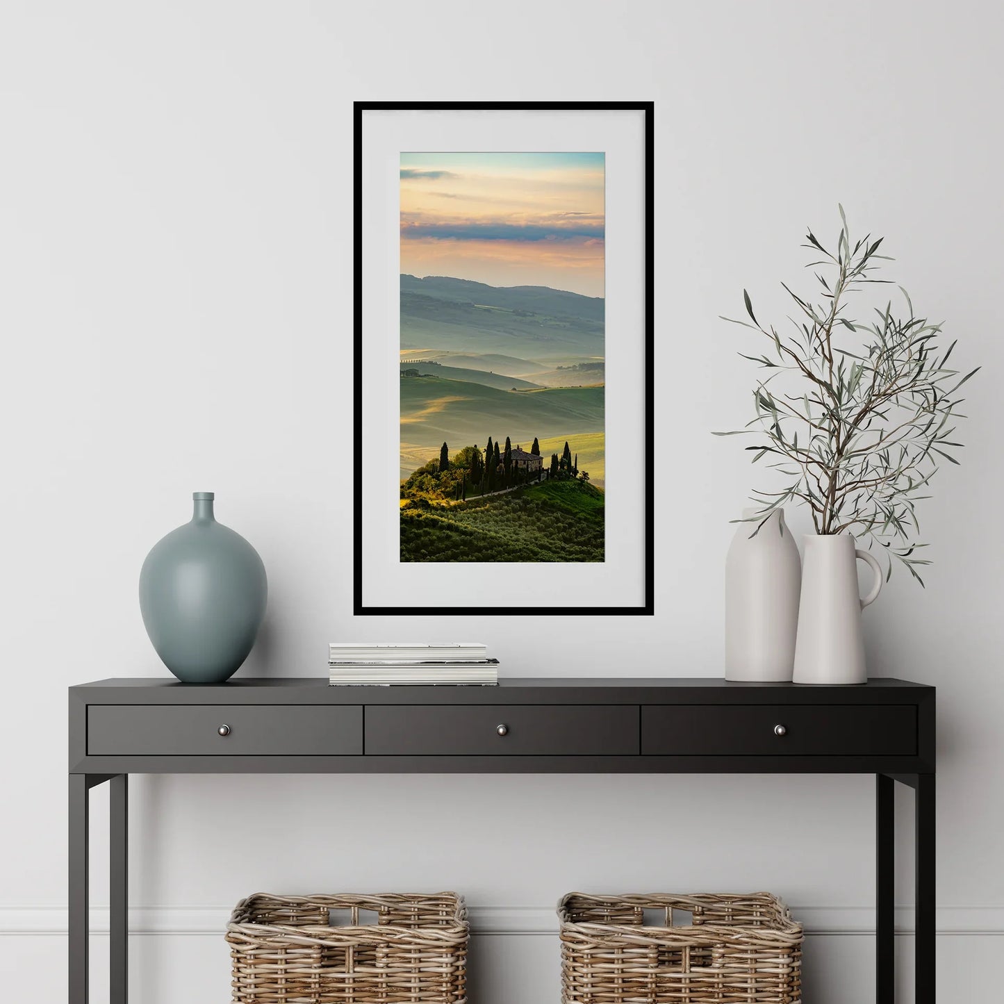 Vertical panoramic print of sunlit rolling hills and a countryside villa at sunrise, framed in black and styled above a console table with decorative vases.