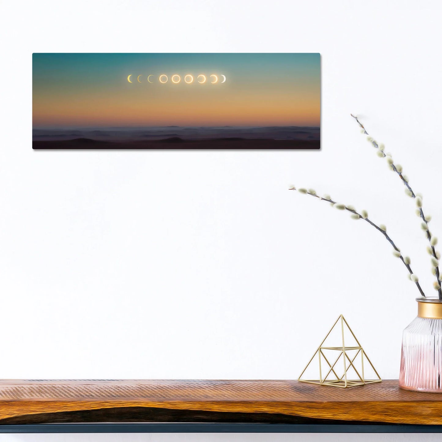 Panoramic print featuring moon phases across a gradient sunset sky above layered mountain silhouettes, mounted on a white wall above a wooden console table.