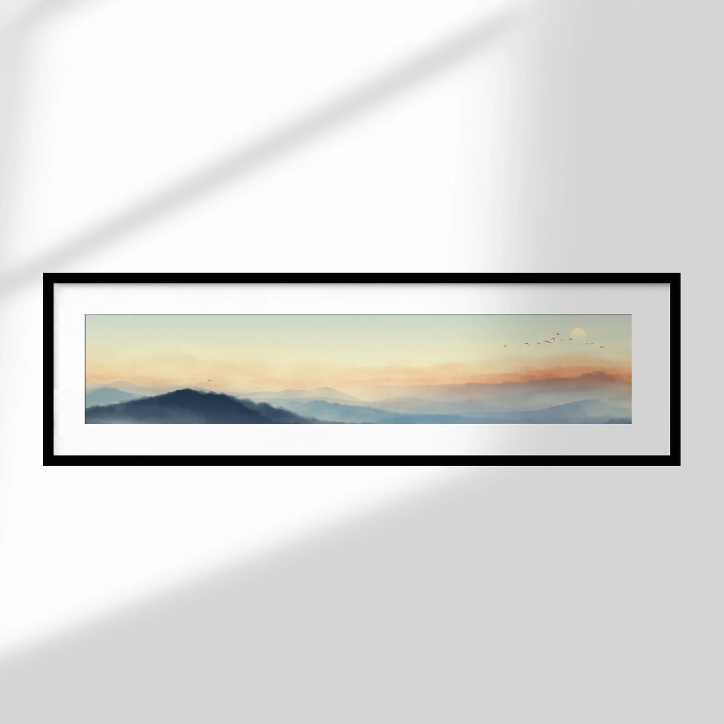 Framed panoramic print with white mat and black frame displaying a soft mountain landscape at sunrise, hung on a neutral wall.