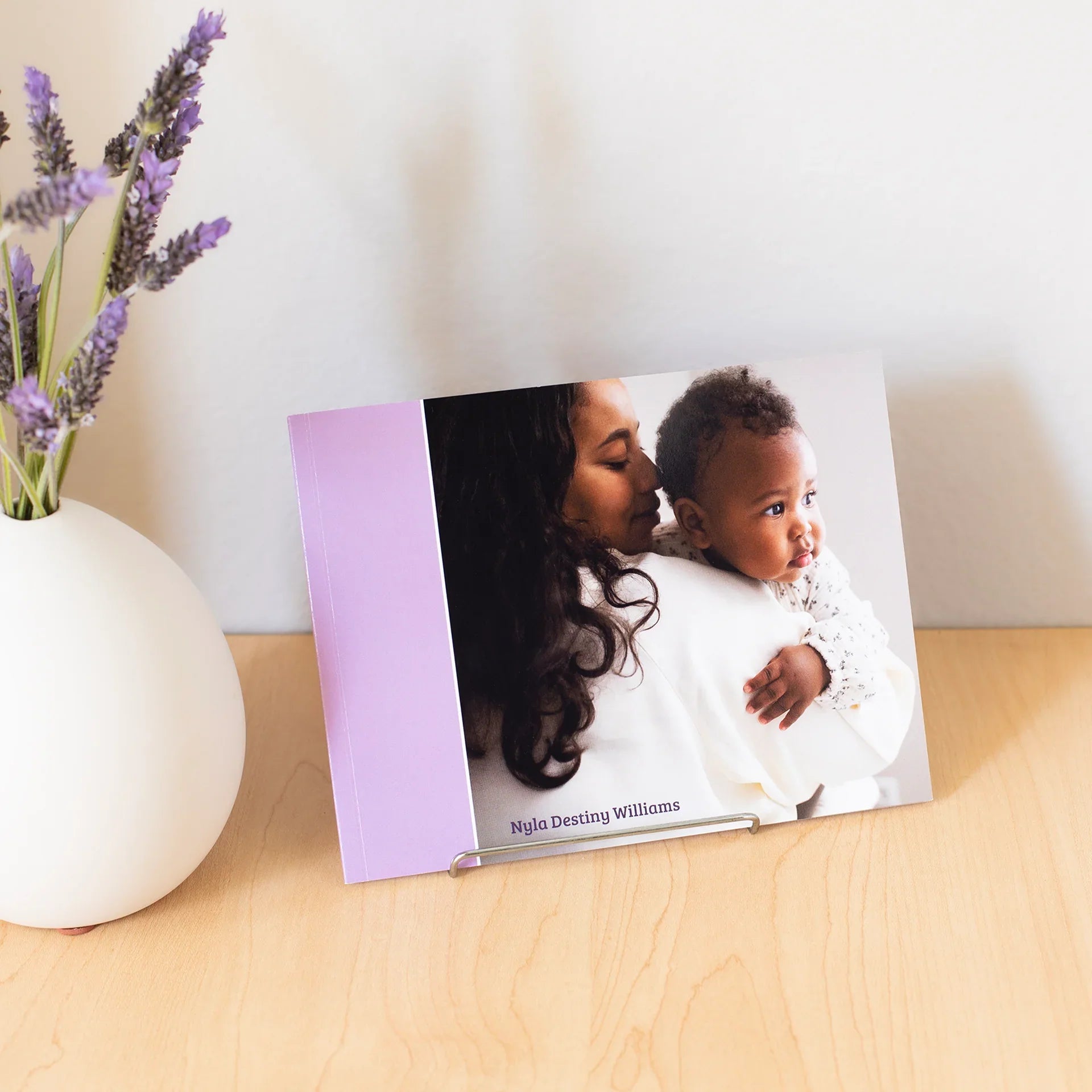 Softcover photo book displayed on a stand, featuring a cover photo of an adult holding a baby, with a lavender spine and name text