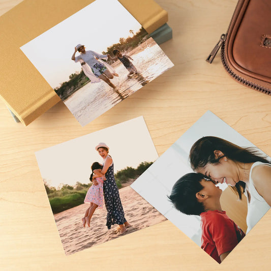 Three glossy photo prints of a mother and child at the beach, arranged on a light wood surface near a fabric-covered journal