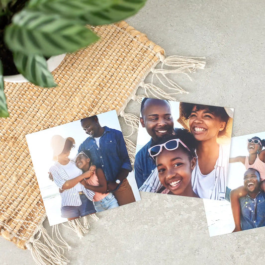 Three glossy family photo prints on a woven mat, featuring parents and child smiling together outdoors in bright natural sunlight.