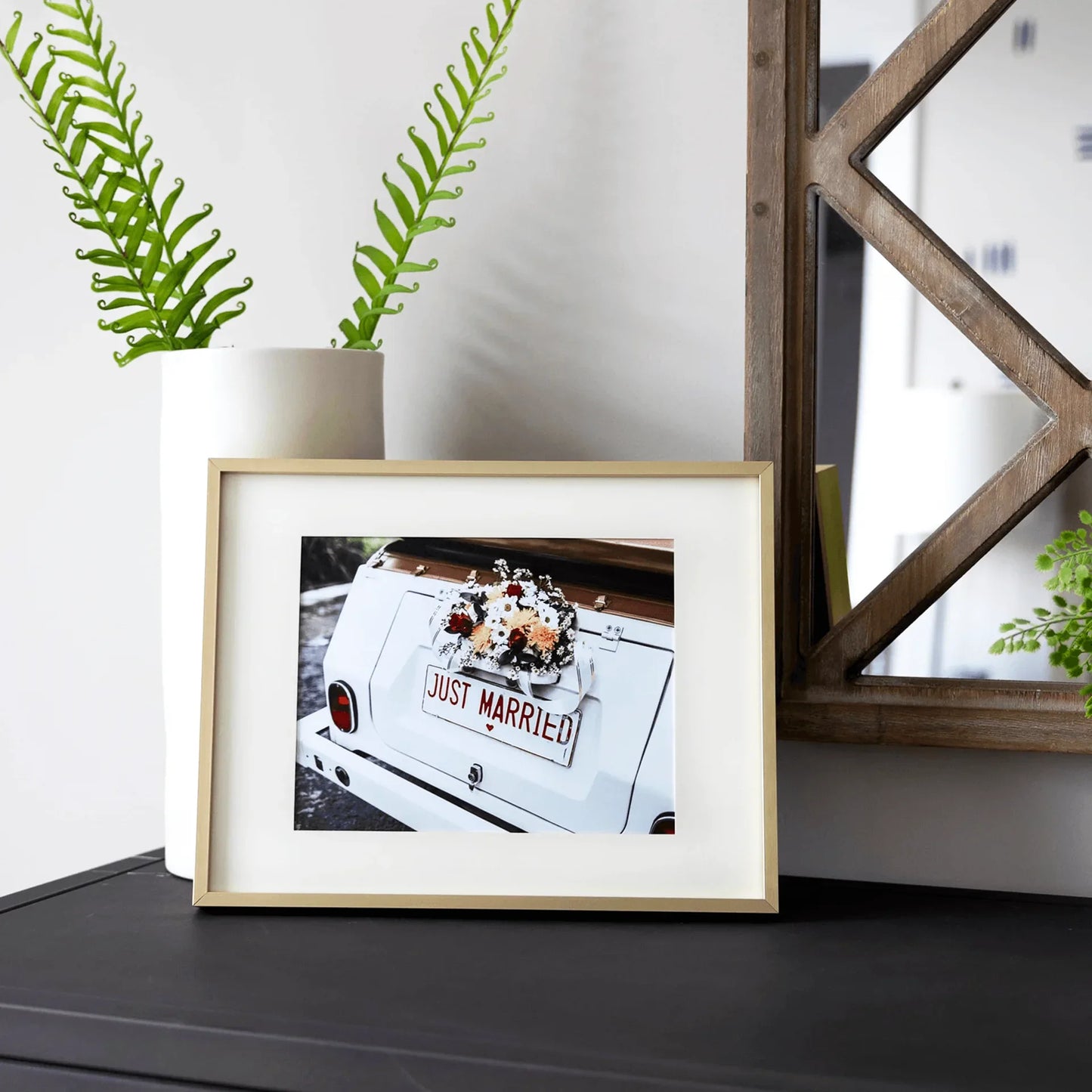 Gold framed horizontal photo print of a vintage white car with “Just Married” sign and floral arrangement, displayed on a dark console table beside a plant.