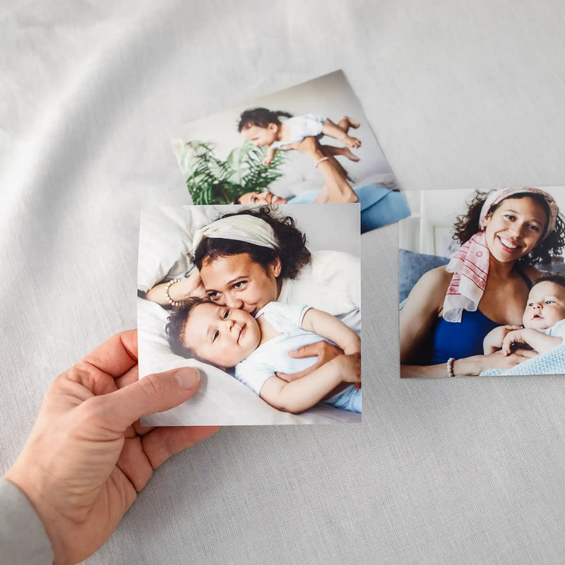 Hand holding a square photo print of a mother cuddling a baby, with two additional square prints arranged on a light fabric surface.