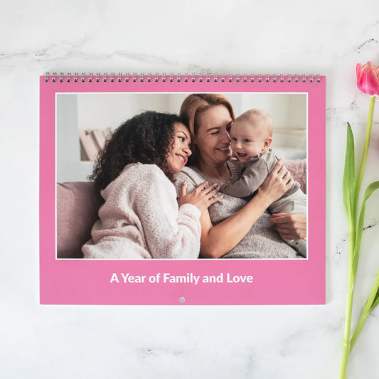 A horizontal wall calendar showing two adults and a baby sitting together on a couch, spiral binding at top and monthly calendar grid below.