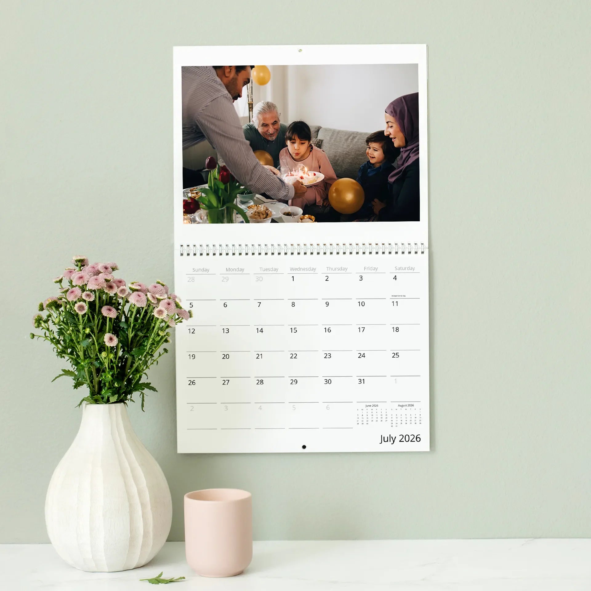 A wall calendar featuring a family gathered around a birthday cake indoors, spiral-bound with a July 2026 monthly calendar grid beneath the photo.