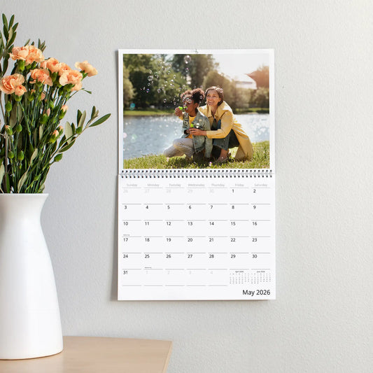 Wall calendar hanging on light wall displaying a color photo of a person and child blowing bubbles by a lake, with monthly grid below.