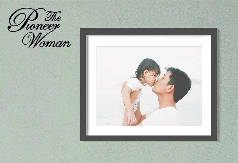 Framed photo print of a parent lifting a child and touching noses displayed on a wall with mat and frame, with “The Pioneer Woman” text.