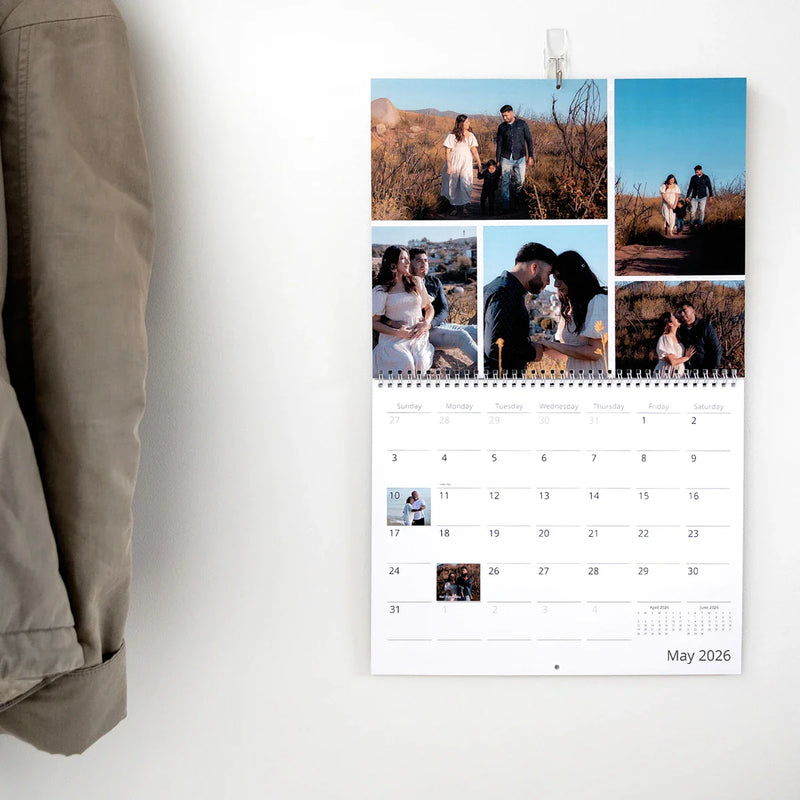 Wall-mounted spiral-bound calendar open to May 2026, featuring a family photo collage at the top and a monthly date grid below on a white wall.